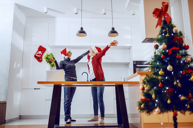 Überglückliche ältere Frau und ihre schwangere Tochter feiern neues Jahr. Beide haben Weihnachtsmützen auf dem Kopf und stehen in der Küche. Silvester.