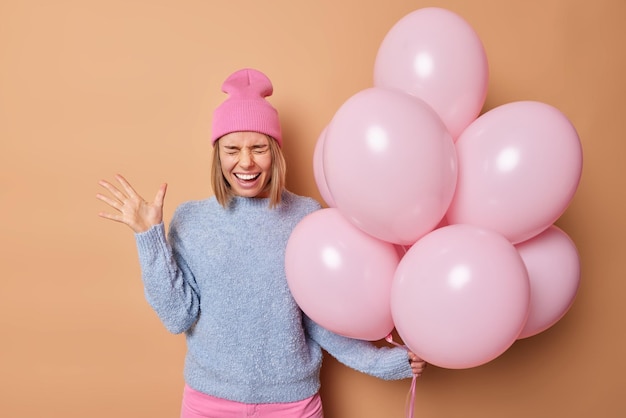 Überglückliche junge Europäerin hält die Handfläche erhoben und ruft laut aus. Sie hält einen Haufen aufgeblasener Luftballons und trägt Hut und Pullover isoliert auf beigem Hintergrund. Menschen Feiertage und Feierkonzept