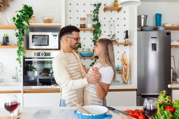 Überglückliche junge Familie, die sich in der modernen Küche amüsiert, tanzt und lacht, glückliche junge Ehefrau und Ehemann, die sich zu Lieblingsmusik bewegen und die Freizeit beim gemeinsamen Abendessen genießen, romantisches Date