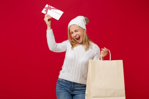 Überglückliche junge Frau im Pullover, Hut isoliert auf rotem Hintergrund. Gesunder Modelebensstil, Konzept der kalten Jahreszeit. Mock-up-Kopienbereich. Halten Sie Paketbeutel mit Einkäufen nach dem Einkauf, Geschenkgutschein.