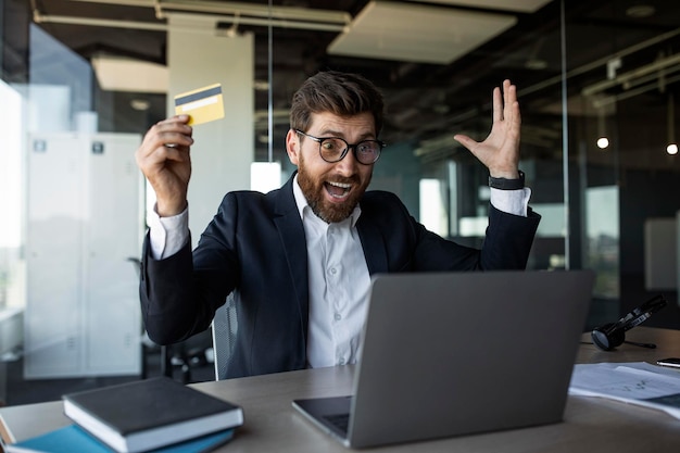 Überglücklicher Geschäftsmann mittleren Alters mit offenem Mund, der schreiend Kreditkarte hält und Laptop im Büro anschaut