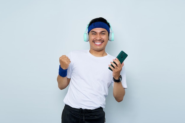 Überglücklicher junger asiatischer Mann sportlicher Fitnesstrainer mit blauem Stirnband und weißem T-Shirt Hören Sie Musik mit Kopfhörern, die Erfolg feiern und Handy auf weißem Hintergrund halten