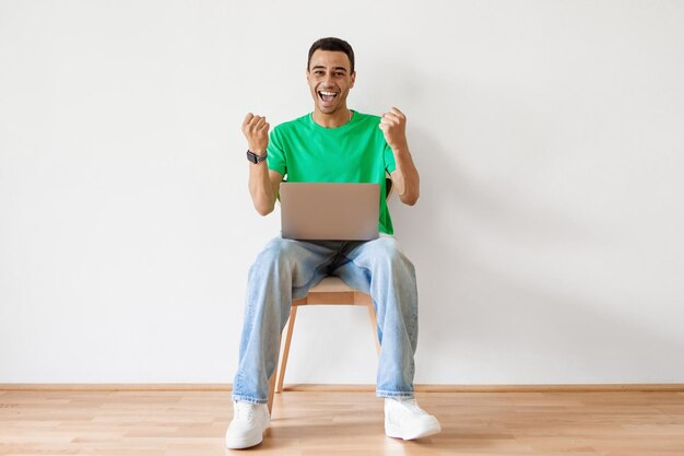 Überglücklicher Mann aus dem Nahen Osten, der mit Laptop auf einem Stuhl sitzt und eine JA-Geste gegen die weiße Studiowand macht