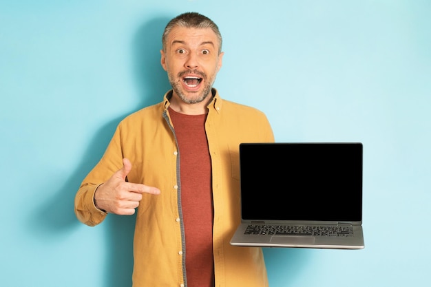 Überglücklicher Mann mittleren Alters, der auf leeren Laptop-Bildschirm mit Attrappe für Website-Design auf blauem Studiohintergrund zeigt