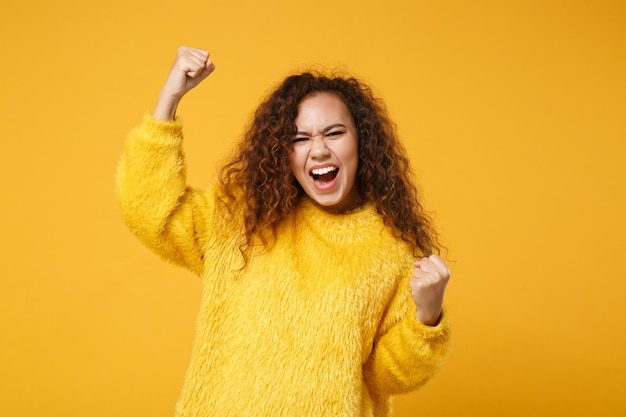 Überglückliches junges afrikanisch-amerikanisches Mädchen im Pelzpullover posiert isoliert auf gelb-orangefarbenem Hintergrund im Studio. Menschen aufrichtige Emotionen Lifestyle-Konzept. Mock-up-Kopienbereich. Fäuste ballen wie Sieger.