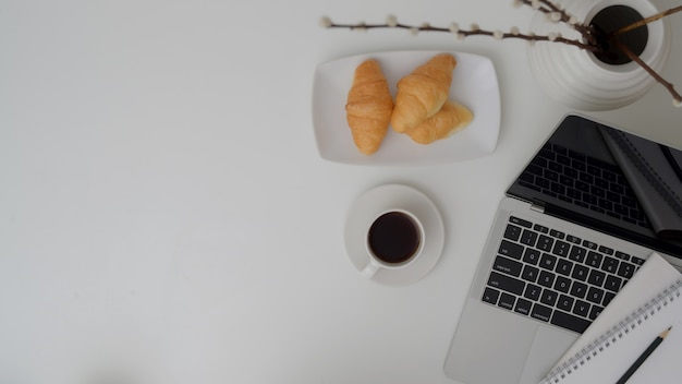 Überkopfaufnahme des Büroraums mit Laptop, Kaffeetasse, Croissant und Vase auf weißem Tisch