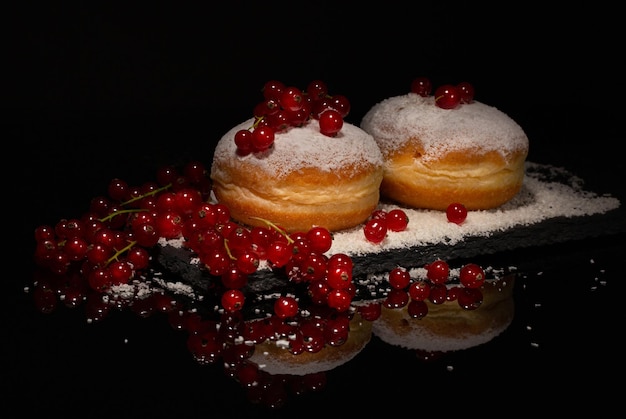 Berliner Deutsche süße Donuts, bestreut mit Puderzucker, gefüllt mit Fruchtmarmelade, umgeben von reifen Beeren auf einem schwarzen Schieferbrett auf schwarzem Hintergrund mit Reflektion