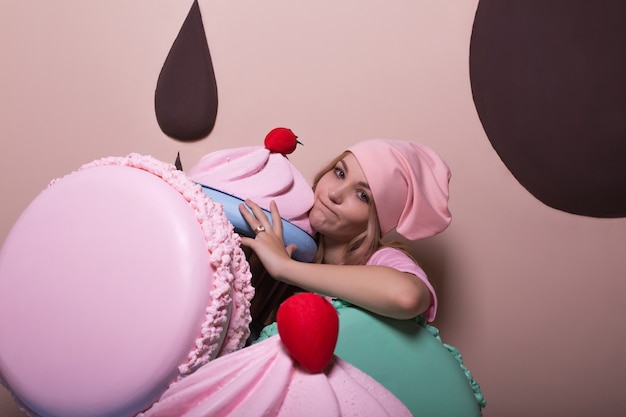 Übermäßiges Essen der blonden Frau mit rosa Mütze und T-Shirt, die Spaß mit großen Makronen und Cupcakes im Studio hat