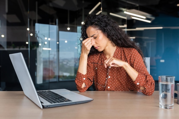 Übermüdete hispanische Frau bei der Arbeit Geschäftsfrau mit Brille, die die Hände auf dem Kopf hält und schwere Schmerzen hat