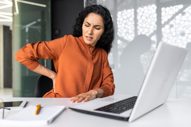 Übermüdete hispanische Frau, die spät arbeitet, Geschäftsfrau, die im Inneren starke Rückenschmerzen hat