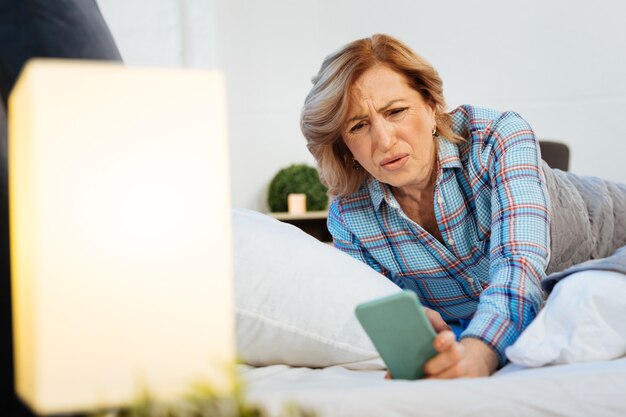 Überprüfungszeit. Verwirrte kurzhaarige erwachsene Frau im hellen Pyjama, die mit unangenehmem Blick auf den Bildschirm ihres Smartphones schaut