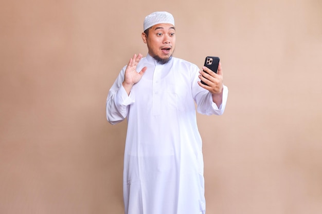 Überraschender muslimischer Mann schaut auf ein Smartphone, das auf einem beigen Hintergrund isoliert ist