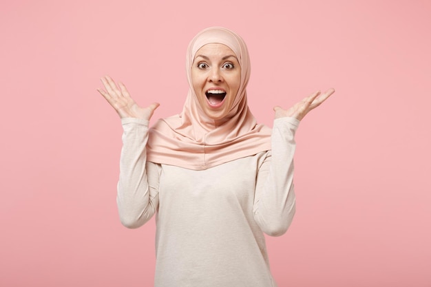 Überrascht erstaunte junge arabische muslimische Frau in Hijab-leichter Kleidung, die isoliert auf rosafarbenem Hintergrund posiert. Menschen religiösen Islam Lifestyle-Konzept. Mock-up-Kopienbereich. Mund offen halten und Hände ausbreiten.