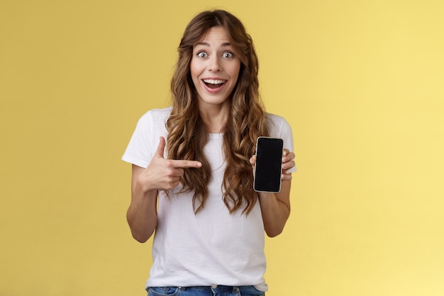 Überrascht glückliches glückliches Mädchen, das die Online-Internet-Lotterie gewinnt und im Allgemeinen lächelt, hält das Smartphone, das auf einen leeren Handy-Bildschirm zeigt, der das Display zeigt, das aufgeregt und enthusiastisch auf gelbem Hintergrund grinst