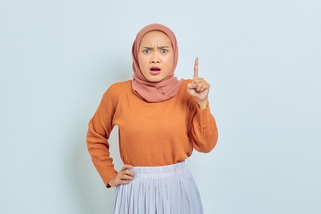 Überrascht junge asiatische Muslimin in braunem Pullover, die mit dem Finger isoliert auf weißem Hintergrund auf Kopierraum zeigt Muslimisches Lifestyle-Konzept