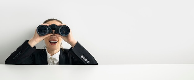 Überrascht junge Frau, die durch ein Fernglas schaut, das hinter einem weißen Pappbanner hervorschaut