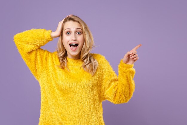 Überrascht junge Frau Mädchen im gelben Pelzpullover posiert isoliert auf pastellviolettem Hintergrund im Studio. People-Lifestyle-Konzept. Mock-up-Kopienbereich. Zeigen Sie mit dem Zeigefinger nach oben und legen Sie die Hand auf den Kopf.