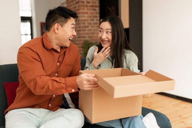 Überrascht koreanisches Paar beim Auspacken von Paketen nach erfolgreichem Einkaufen auf dem Sofa im Wohnzimmer zu Hause