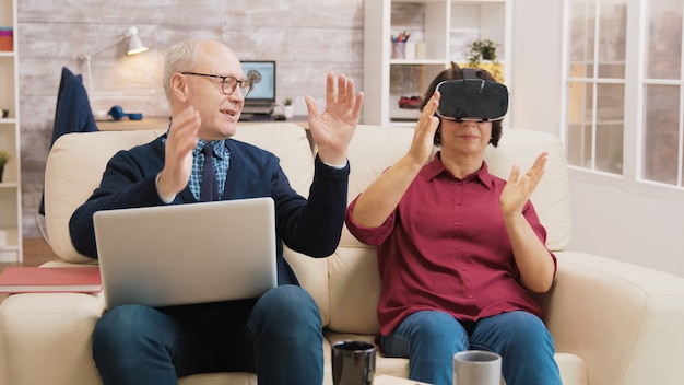 Überraschte ältere Frau, während sie mit ihrem Mann neben ihr mit dem Laptop eine Virtual-Reality-Brille auf dem Sofa verwendet.