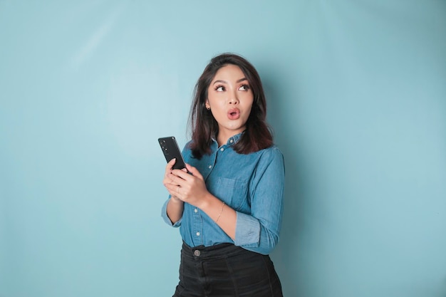 Überraschte asiatische Frau mit blauem Hemd, die auf ihr Smartphone zeigt, das durch blauen Hintergrund isoliert ist