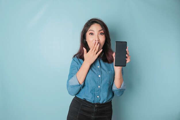 Überraschte asiatische Frau mit blauem Hemd, die auf ihr Smartphone zeigt, das durch blauen Hintergrund isoliert ist