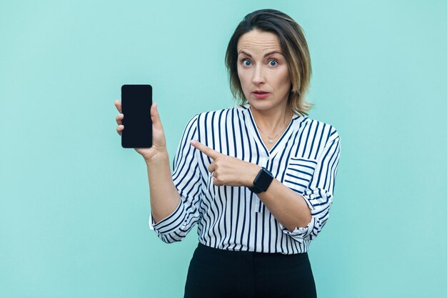 Überraschte blonde Geschäftsfrau, die mit dem Finger auf das Smartphone zeigt und mit geschütteltem Gesicht in die Kamera schaut