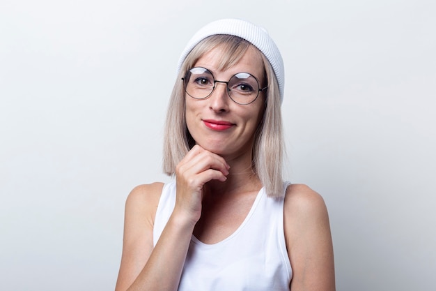 Überraschte blonde junge Frau mit Brille, weißer Hut auf hellem Hintergrund.