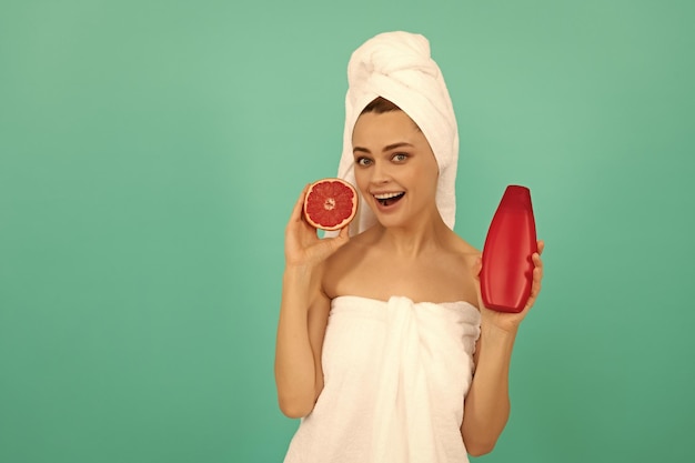 Überraschte Dame im Handtuch mit Grapefruit-Shampoo-Flasche auf blauem Hintergrund