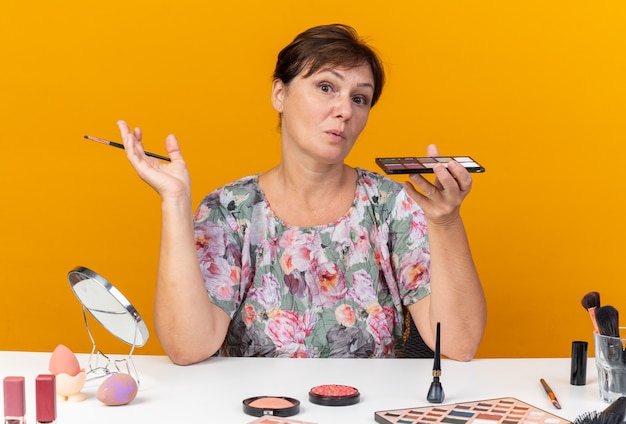 Überraschte erwachsene kaukasische Frau, die am Tisch mit Make-up-Tools sitzt und Lidschatten-Palette und Make-up-Pinsel hält