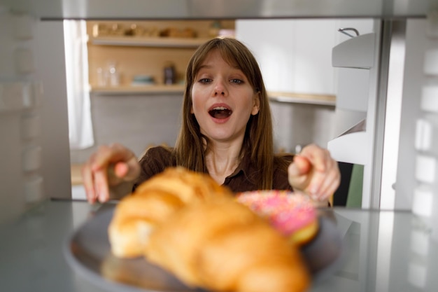 Überraschte Frau, die Teller mit Croissants und Donut aus dem Kühlschrank nimmt