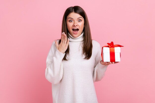 Überraschte Frau mit dunklem Haar, die eingewickelte Geschenkbox hält und mit Erstaunen in die Kamera blickt und einen weißen Pullover im lässigen Stil trägt Studioaufnahme isoliert auf rosa Hintergrund