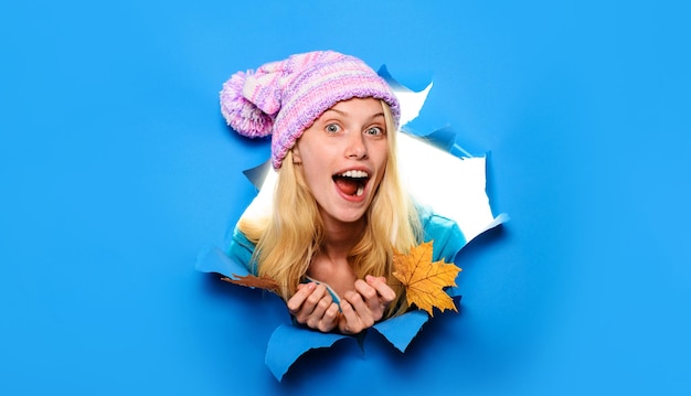 Überraschte Frau mit gestricktem Hut mit Herbstblättern, die durch ein Papierloch schaut