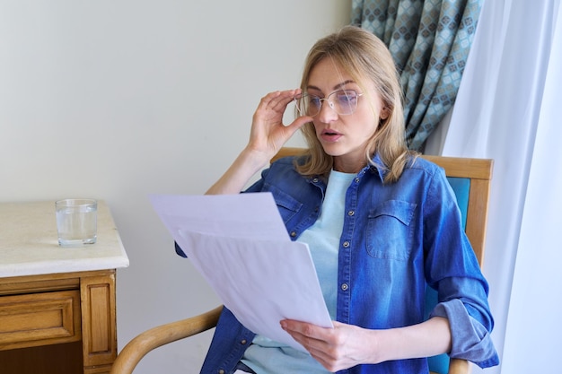 Überraschte Frau mittleren Alters mit Brille, die zu Hause auf einem Stuhl sitzt und Papiere liest