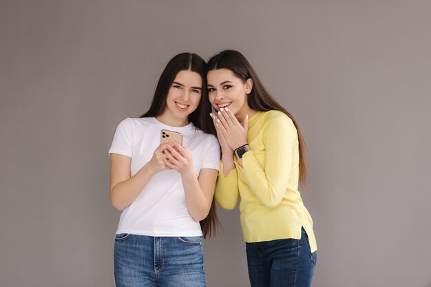 Überraschte Frauen schauen auf das Telefon und schließen den Mund mit den Händen attraktiver junger Frauen im Studio auf Grau