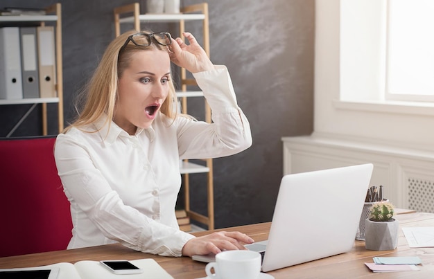 Überraschte Geschäftsfrau mit Brille, die am Laptop arbeitet, während sie an ihrem Arbeitsplatz im Büro sitzt