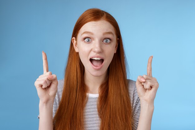 Überraschte glückliche begeisterte junge rothaarige Frau reagierte beeindruckt obere hängende Promo zeigt nach oben Zeigefinger Tropfen Kiefer amüsierten Blick begeistert Kamera erzählt von fantastischen Angebot blauen Hintergrund