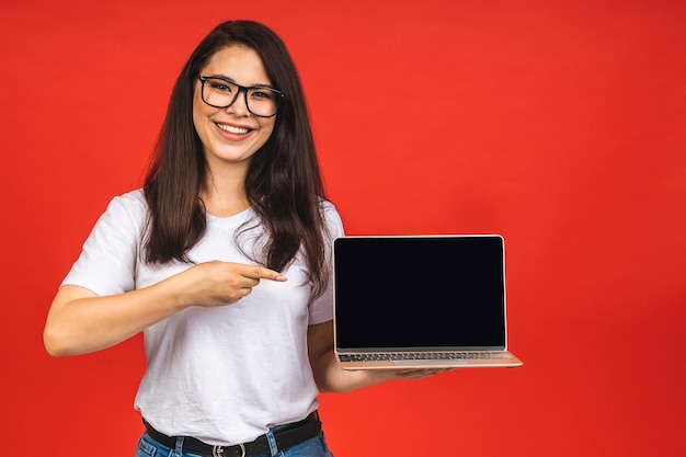 Überraschte, glückliche, brünette Frau in legerer Kleidung, die einen leeren Laptop-Computerbildschirm zeigt, während sie isoliert auf rotem Hintergrund in die Kamera schaut