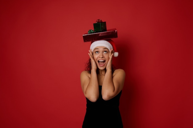 Überraschte hübsche Frau in Weihnachtsmütze und schwarzem Abendkleid, die jubelnd zu Weihnachtsgeschenkboxen auf ihrem Kopf aufschaut, ihre Hände auf ihre Wangen nimmt und Glück ausdrückt, einzeln auf rotem Hintergrund