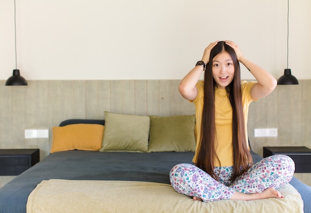 Überraschte junge asiatische Frau am Bett mit den Händen auf Kopf