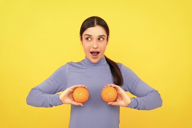 Überraschte junge Frau, die orange Zitrusfrüchte auf natürlichem Lebensmittel des gelben Hintergrundes hält