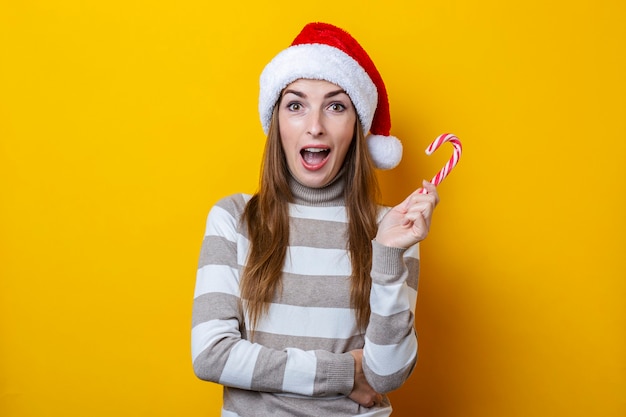 Überraschte junge Frau im Weihnachtsmann-Hut mit Weihnachtssüßigkeit auf gelbem Hintergrund.