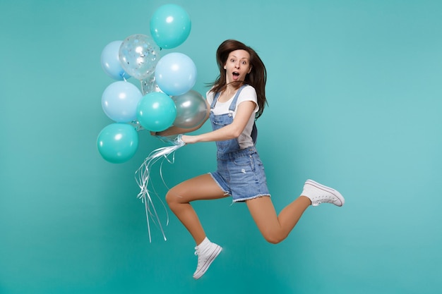 Überraschte junge Frau in Denim-Kleidung, die den Mund offen hält, hoch springt, feiert, bunte Luftballons auf blauem türkisfarbenem Hintergrund hält. Geburtstagsfeier, Menschen Emotionen Konzept.