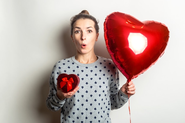 Überraschte junge Frau in einem Pullover hält ein Geschenk und einen Herzluftballon auf einem hellen Hintergrund. Valentinstag Konzept. Banner.