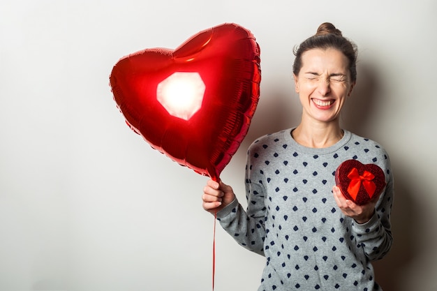 Überraschte junge Frau in einem Pullover hält einen Poarok für und Herz Luftballon und freut sich über das Geschenk auf einem hellen Hintergrund. Valentinstag Konzept. Banner.