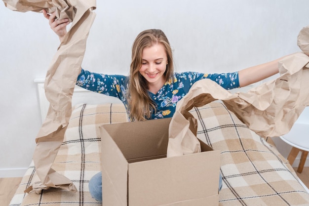 Überraschte junge Frau sitzt auf dem Bett und packt eine Kartonbox zu Hause aus und liefert ein Paket.