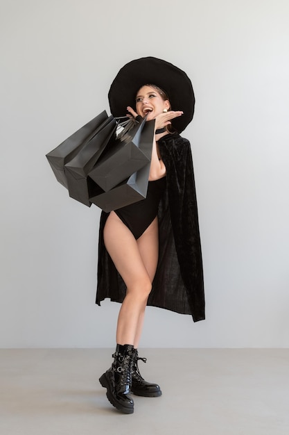Überraschte junge glückliche sexy Frau in schwarzem Hexenhut mit Einkaufstaschen nach erfolgreichem Einkaufen Halloween-Verkauf Rabatte