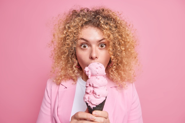 Überraschte lockige junge Frau leckt Eis isst Junk-Food isst appetitliches Sommerdessert in stilvollen Outfit-Posen gegen rosa Wand. Schöne europäische Frau beißt süßen Snack