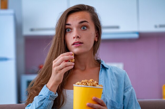 Überraschte schockierte ängstliche Frau, die knuspriges Karamellpopcorn beim Anschauen des Horrorfilms zu Hause isst. Popcorn-Film