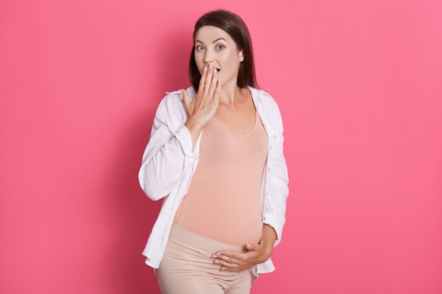 Überraschte schwangere Frau, die mit geöffnetem Mund aufwirft, Mund mit Handfläche bedeckt, isoliert über rosa Hintergrund aufwirft, ihren Bauch berührt, Leggins und Hemd trägt.