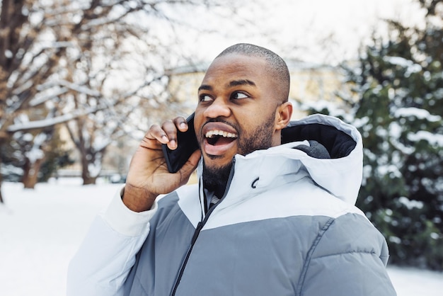Überraschter afrikanischer Mann, der am Telefon spricht, lächelnder Winterspaziergang im Freien